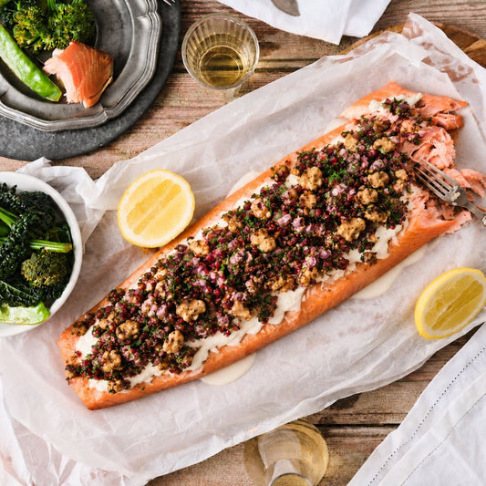 A baked salmon fillet topped with a tahini yoghurt and walnut tarrator, served on parchment paper with lemon wedges and a fork on a rustic wooden table.