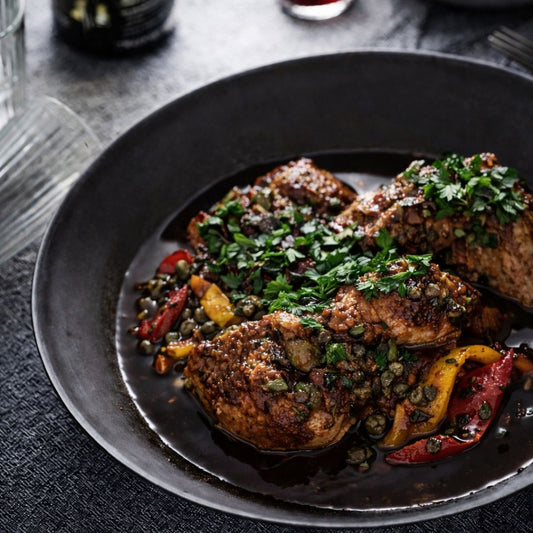 Plated dish of chicken trasteverina with roasted red peppers, baby capers, jus gras in a dark bowl
