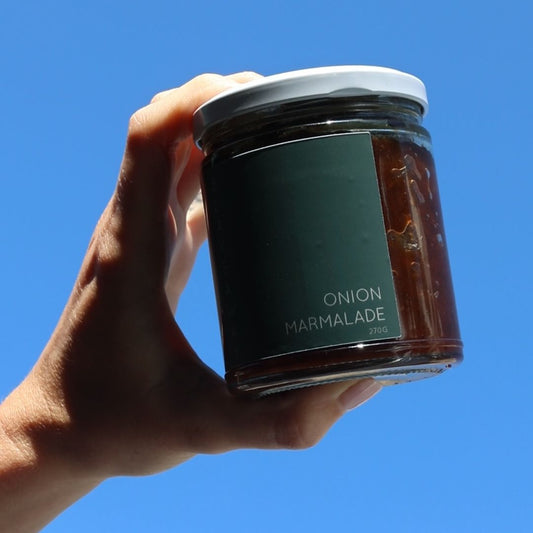 A hand holding a glass jar of onion marmalade against a clear blue sky. The jar has a dark green label with white text and a white lid.