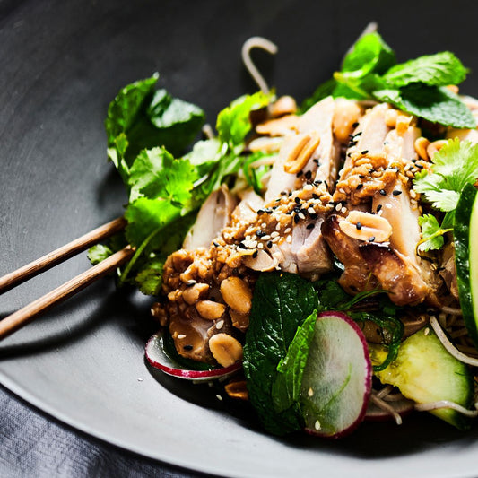 Soba noodles with teriyaki chicken, vegetables, and sesame seeds on a dark plate with chopsticks.