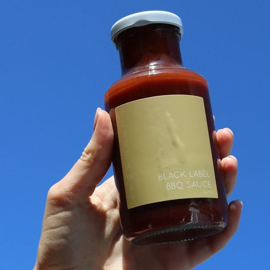 A hand holding a bottle of Black Label BBQ Sauce against a clear blue sky background.