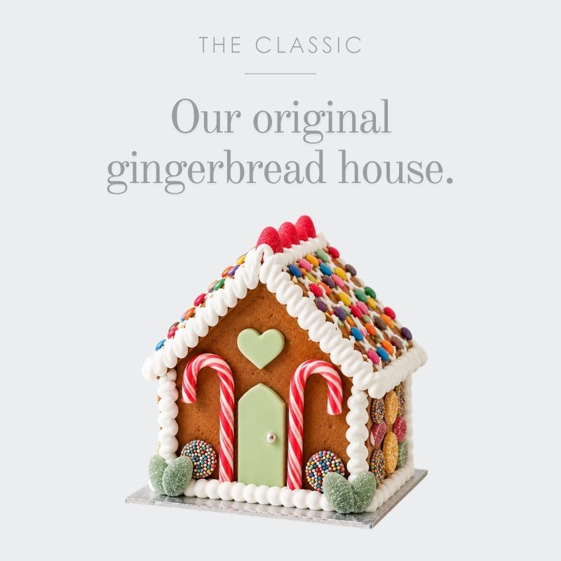 A decorated gingerbread house cake with white piped icing borders, colourful candy sprinkles on the roof, candy cane doors, a light green heart-shaped window, and a light green door, shown below the text 'THE CLASSIC Our original gingerbread house.' on a light grey background.