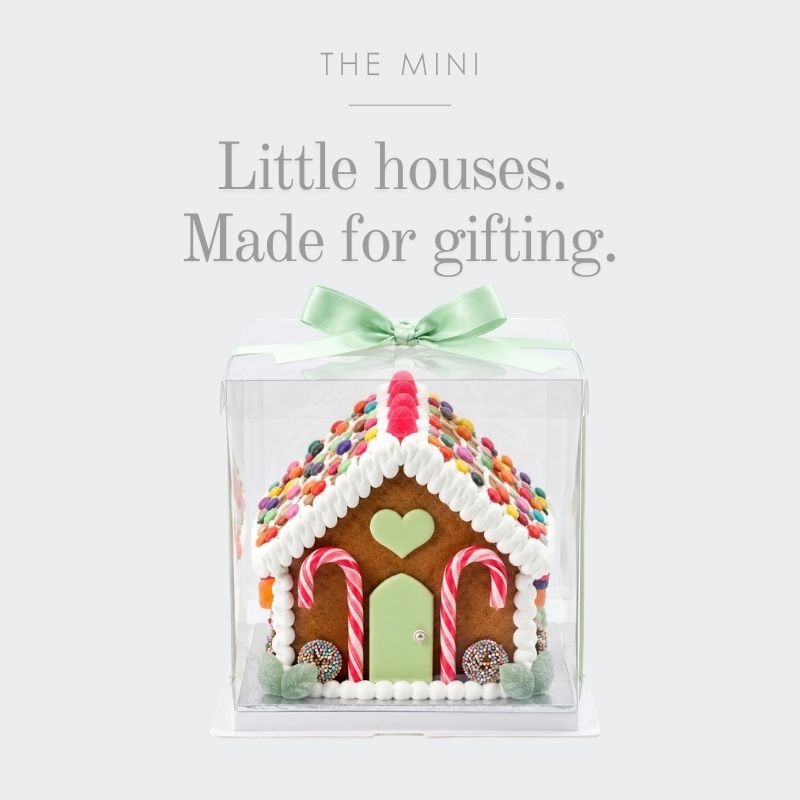 A gingerbread house gift box decoration set inside a clear square plastic container, tied with a mint green ribbon. The house is decorated with white icing, colourful sprinkles, candy canes, and a green door.