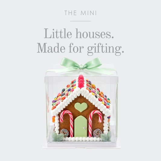 A gingerbread house gift box decoration set inside a clear square plastic container, tied with a mint green ribbon. The house is decorated with white icing, colourful sprinkles, candy canes, and a green door.