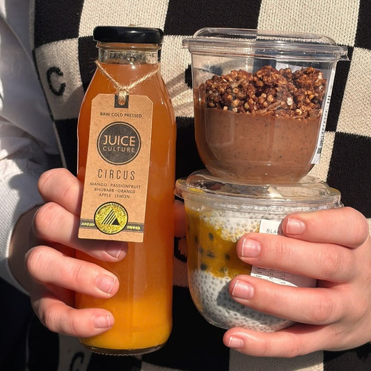 A person holds two food items: a bottle of Juice Culture Circus mango passionfruit rhubarb orange juice, and a stacked dessert of chocolate pudding topped with granola and a clear cup of chia pudding with passionfruit seeds.