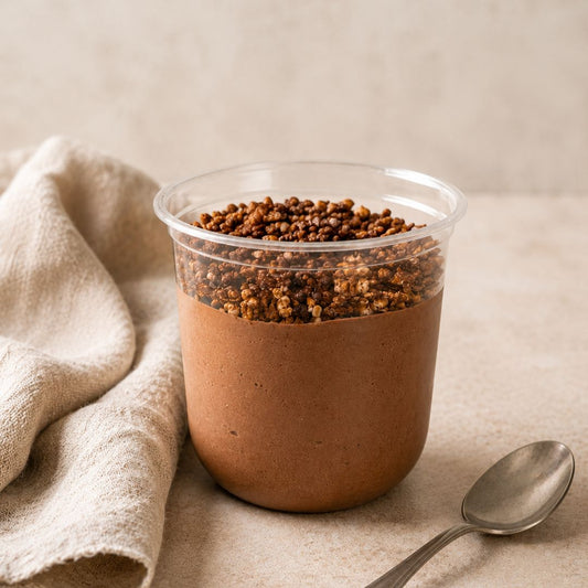 A clear plastic cup filled with dark chocolate protein mousse and topped with crumbled quinoa, set on a beige surface with a folded linen cloth and a spoon nearby.