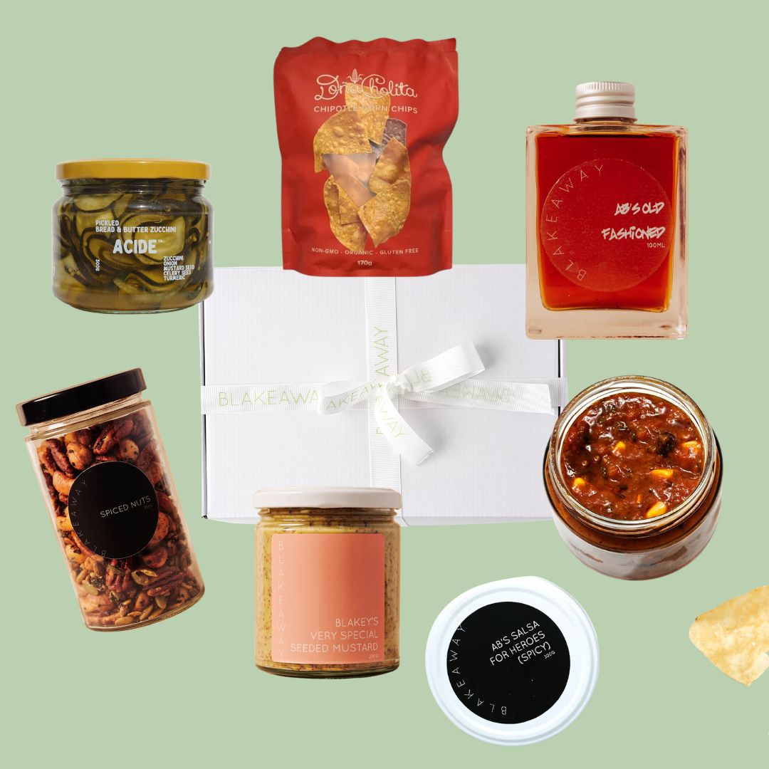 A curated gift basket arrangement featuring a white gift box tied with a white ribbon, surrounded by assorted gourmet snacks and drinks: a jar of pickled zucchinis, a red bag of chips, a bottle of old fashioned, a jar of spiced nuts, a jar of salsa, a small jar of special seeded mustard.