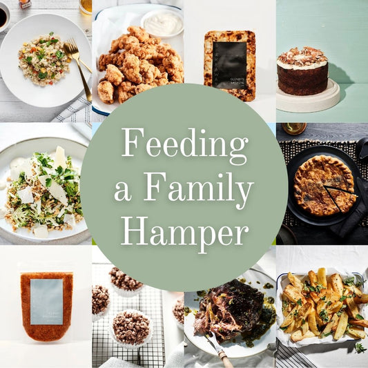 A collage of food items and serving dishes arranged around a large muted green circle with the text 'Feeding a Family Hamper' in white. Visible food includes a cabbage & broccoli slaw, popcorn chicken, carrot cake, chocolate crackles, chicken & leek pie, lemon greek roast potatoes, vegetarian fried rice.