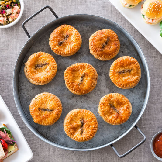Golden-brown flaky pies arranged on a round grey metal serving tray, photographed from above on a light gray tabletop with partial side dishes visible around the edges.