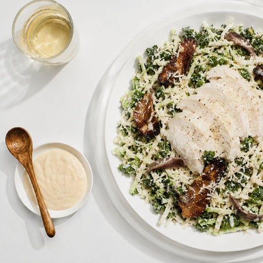 A plated salad featuring sliced white chicken breast over a bed of shredded cabbage and grated cheese, with a glass of white wine, a small bowl of creamy dressing, and a wooden spoon on a white surface.