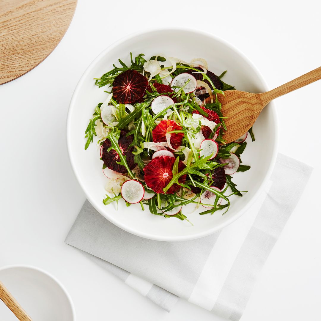 White bowl with a colourful rocket, baby fennel, radish and orange salad; with a wooden spoon on a white background