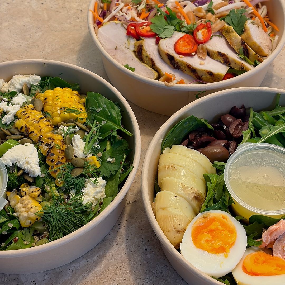 Three bowls of prepared salads and sliced grilled chicken on a light stone surface. One bowl contains mixed greens, corn, feta cheese, and olives; another has sliced chicken, red peppers, and shredded carrots; the third shows greens, hard-boiled eggs, olives, and a small clear dressing cup.