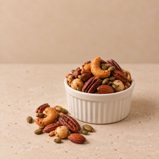 A white ribbed ramekin filled with mixed nuts and seeds, including cashews, almonds, pecans, and pumpkin seeds, with a few scattered on a beige surface against a matching beige background.
