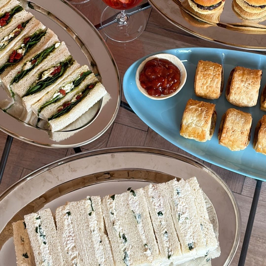 An overhead view of an elegant catered food spread on a wooden table, featuring sliced sandwiches on gold-rimmed plates, golden-brown sausage rolls on a light blue platter, and a small bowl of tomato relish.