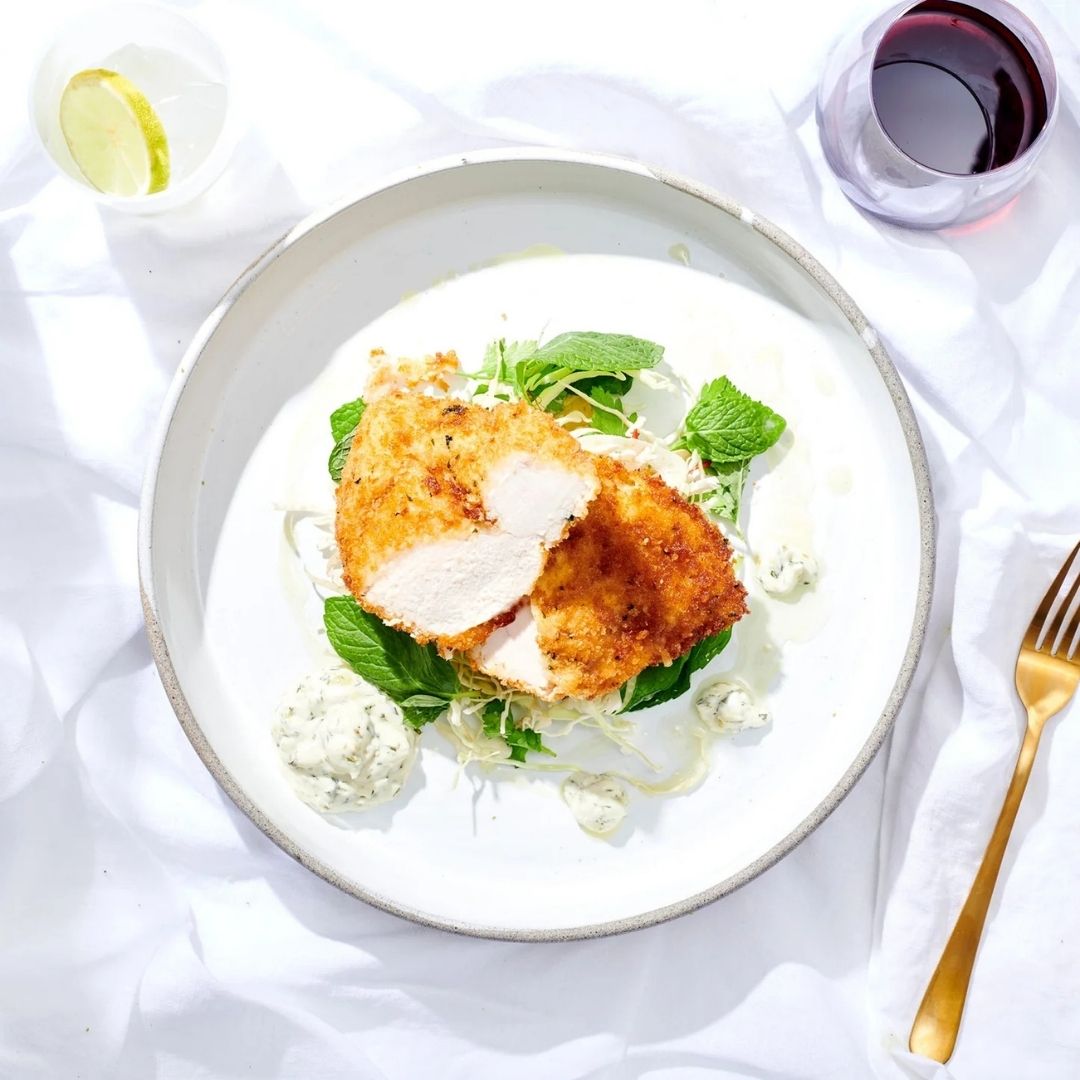 Plated dish of crumbed chicken schnitzel with greens on a white tablecloth with a glass of red wine.