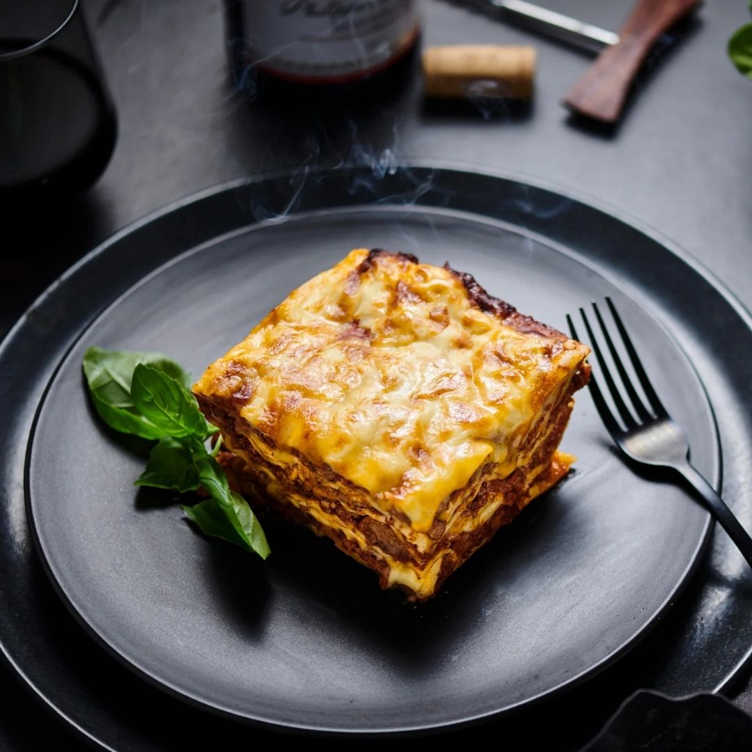 Glenby's lasagna on a black plate with a fork, on a dark surface.